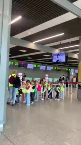 Here is a description of the video:

The video shows a group of children, most likely a school class on an outing, lining up at the check-in counters of an airport. They are accompanied by at least one adult. The children are wearing bright yellow reflective vests and carrying backpacks. They are waiting in front of check-in counters labeled with numbers 26 through 32. Above the counters are digital screens displaying flight information and the word "Welcome." There is a screen in the background displaying the word "DORMITORIUM." The airport interior appears modern, with a high ceiling and a sleek design. The atmosphere seems calm, as people are waiting in an orderly fashion.