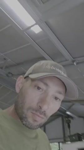 A man wearing a light green t-shirt and a light-colored baseball cap stands outside. The cap has "Flyboard" written on it. The man has a trimmed beard and is looking directly at the camera. Behind him is a gray concrete driveway with two vehicles parked in the distance, as well as a cluster of trees and bushes. A gray garbage can is partially visible to the right. The man appears to be filming himself. The video appears to be slightly overexposed.