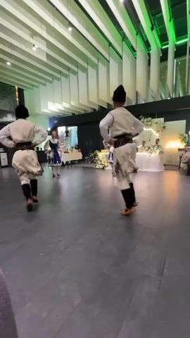 The video shows four people dancing in what appears to be an indoor event space. Three individuals are dressed in traditional folk costumes. Two men in the foreground are wearing white tunics with dark sashes, dark pants, and dark hats. They are performing synchronized dance moves, including jumps and kicks. A woman in the background is wearing a blue and white folk dress. She is dancing with another woman in a similar outfit. In the background, there is a table with floral arrangements, suggesting a wedding or formal gathering. The room has a modern aesthetic with high ceilings, exposed beams, and green accent lighting. The floor is dark and reflective.