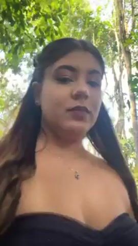 The video shows three women walking in a forested area. The first woman, who seems to be holding the camera, is wearing a dark blue, strapless dress. Behind her is another woman in a dark red, strapless dress with a bow in the back. A third woman, wearing a yellow, strapless dress, walks in front of the other two. The area they are walking through appears to be natural, with trees and foliage on either side of a sandy path.