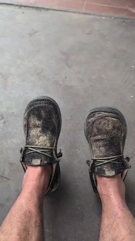 The video shows a pair of dirty, well-worn casual shoes on someone's feet, viewed from above. The shoes are a dark color with lighter, muddy or dusty patches across the tops. The laces are visible and appear slightly frayed. The background is a concrete floor with some scattered debris and a hint of a tiled area. In the final frame, a white athletic shoe with red accents enters the frame from the left, as if someone is walking by. The person wearing the casual shoes remains stationary throughout the clip.