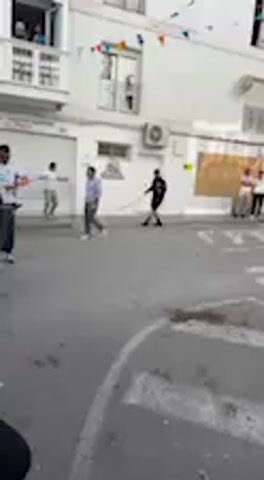 The video shows a street scene in what appears to be a European town, confirmed by context as Vejer de la Frontera, Spain. Whitewashed buildings with balconies, windows, and air conditioning units line the narrow street. Colorful bunting is strung across the street, suggesting a festive occasion.

Several people are present. On the right side of the street, a group of adults is standing on the sidewalk, observing an activity. On the left, a person runs across the street and then crouches down near the base of a building. Further down the street on the left, more individuals are visible in the distance. Another person appears to be holding a long stick or pole. The street itself has painted white markings, including what looks like a crosswalk and a turn arrow. The weather appears sunny and clear, typical of daytime. The overall atmosphere suggests a community gathering or a traditional street game is taking place. There is no clearly visible text in the scene.