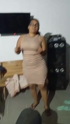 A woman is dancing in a room. She is wearing a striped dress and glasses. There is a television and speakers in the background. There is also a couch and some flip flops.