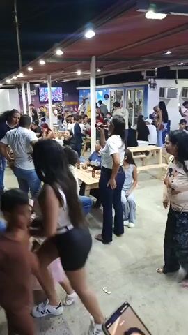 The video shows a crowded outdoor event at night. People are sitting at picnic tables, drinking and talking. There is a stage in the background with a large screen displaying visuals. Some people are walking around, including a child in a tutu. The atmosphere appears festive and lively.