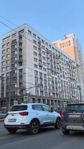 The video depicts a street scene in an urban environment. The foreground is dominated by moving traffic, including a white SUV and a silver car. Pedestrian crosswalks are visible at intersections, and streetlights are present. In the background, tall, multi-story buildings with numerous windows and balconies rise against a clear, blue sky. Some of the buildings are adorned with architectural details such as cornices and columns. Cables and wires are strung overhead. The overall atmosphere is one of a typical, bustling city street.