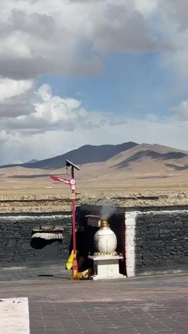 The video captures an outdoor scene in Labrang township, China, showcasing a cultural or religious site under a partially cloudy blue sky during daytime.

In the foreground, a dark-colored paved ground extends towards a low, stone wall constructed from rough-hewn blocks, segmented into black and black-and-white sections. Central to the frame, within a recess of this wall, sits a white, ornate censer or incense burner with a golden top, resting on a square white pedestal. Wisps of smoke steadily rise from the censer, indicating a continuous burning offering or incense.

To the left of the censer, a tall, slender red pole stands upright. It features an ornate, possibly traditional, decorative element at its apex, below which a small solar panel and an attached light or camera device are visible. A piece of yellow fabric is draped at the base of this pole.

The background reveals a vast, arid landscape characterized by gently rolling, treeless mountains under the expansive sky. Between the wall and the distant mountains lies a flat, light-brown plain, typical of high-altitude regions. The overall scene conveys a sense of peaceful adherence to cultural practices amidst a rugged natural environment. No people are visible in the video frame. The audio contains speech in what sounds like Chinese.