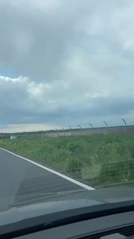 The video shows a view from inside a car driving on an asphalt road.  The camera faces forward, showing the road ahead, which is marked with a solid white line on the left and broken white lines in the center. The landscape to the right of the road features green vegetation and a fence with barbed wire on top in the foreground, with fields stretching into the distance. The sky is partly cloudy, with patches of blue sky visible between the clouds and sun rays shining through. There is audio of people talking.
