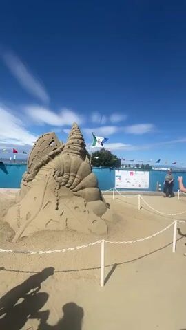 The video shows a sunny day with a clear blue sky and scattered clouds. It appears to be an outdoor event, possibly a sand sculpture festival, set up on a sandy area near a body of water, likely a lake or ocean. Several elaborate sand sculptures are visible, some resembling sea creatures and abstract shapes. People are walking around and viewing the sculptures. In the foreground, a young girl in a black skirt and white top is walking away from the camera, reaching out towards a large sand sculpture. In the background, there are flags, banners, and other attendees. The area is sectioned off with white ropes and posts. The overall atmosphere is that of a public exhibition on a pleasant day.