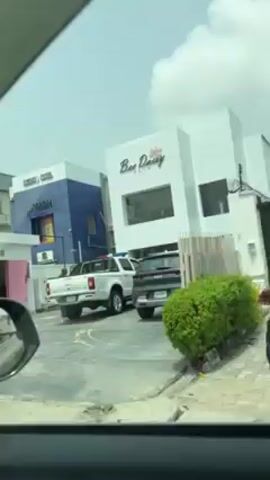 The video shows a view from inside a car, looking out at a commercial street. There are several buildings visible. One has a blue facade and says "GOLD MALL" on its side. Next to it is a white building labeled "Bee Daisy." A white pickup truck with emergency lights on its roof is parked in front of the Bee Daisy building, along with a dark SUV. A green bush is in front of a fence beside the road. The sky is partly cloudy. The car appears to be moving.