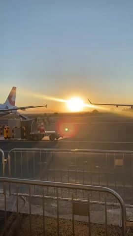 This video captures a serene sunrise at an airport, likely in Zemunik Donji, Croatia. The sun, a brilliant orb, ascends over a hazy horizon, casting long shadows and a warm, golden glow across the tarmac. The scene is dominated by two airplanes, their sleek silhouettes stark against the bright sky. One plane, on the left, is positioned closer to the foreground, its tail fin subtly visible. Another plane, further in the distance on the right, is partially obscured by the sun's glare, with its wing extending towards the viewer.

In the middle ground, several objects suggest airport operations. A baggage cart with orange safety cones is parked near the first plane, indicating ground support activities. A metal barrier stretches across the lower part of the frame, separating the viewer from the operational area of the airport. The clear, pale blue sky suggests fair weather. The overall mood is peaceful and tranquil, evoking the quiet anticipation of a new day at the airport. There is no visible text in the video.