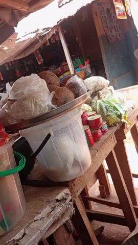 The video shows a market stall with a variety of items for sale. On the wooden table are coconuts, bags of what appear to be processed food, canned goods, and leafy green vegetables.  A clear plastic bucket contains what appears to be a white, wet substance, possibly soaked grains or food.  Another bucket, partially visible, contains packaged foods.  Behind the table, a makeshift roof provides shelter.  Above and behind the table are more shelves and stalls, indicating a bustling market environment. The camera pans across the stall, showing different containers of goods.