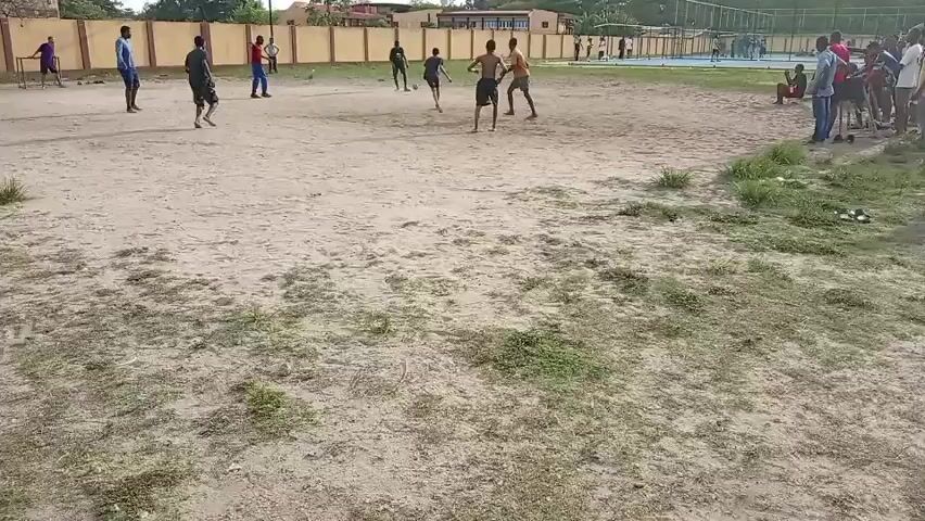 Several people are playing a game of soccer on a sand-covered field. Spectators are standing along the right side of the frame. The field is bordered by a low brick wall and trees in the background. A sports fence and another field are visible in the distance. The lighting is bright, suggesting it is daytime. The ground has patches of grass. The people are wearing casual clothing. The focus is on the players as they maneuver the soccer ball.