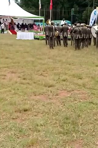The video shows a marching band in uniform moving across a grassy field. The band members are wearing matching dark jackets with light-colored trim, light-colored caps, and dark trousers. They are carrying various musical instruments, including tubas, trombones, and drums. The band is marching away from the camera, towards a large white tent and a raised platform with people seated on it. There are flags and banners visible in the background. The field is a mix of green grass and patches of bare earth. The background includes trees and what appears to be a vehicle. The camera is relatively static.