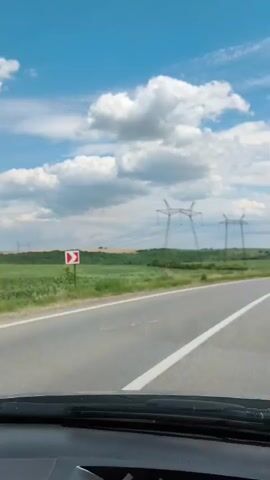 The video shows a view from inside a car, looking out at a rural landscape. A paved road with a white line is visible in the foreground, with the dashboard of the car underneath. Beyond the road, there is a green field of crops. In the background, there are several large power line towers with wires stretching between them. The sky is blue with scattered white clouds. There are also some trees and rolling hills in the distance. The sun appears to be shining.