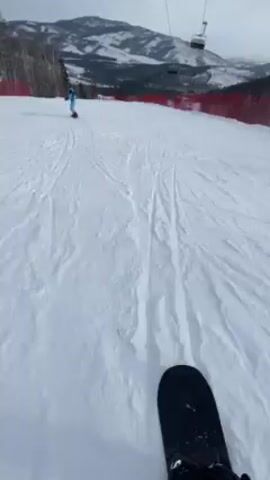 The video shows a first-person view of someone snowboarding down a snow-covered slope on a cloudy day. Another person is snowboarding ahead. A ski lift and mountains are visible in the background. Red fencing borders the side of the slope. The snow is textured with tracks from previous skiers or snowboarders.