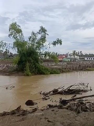 A video shows a muddy river with debris in the foreground. On the far bank, there is a partially constructed bridge with wooden posts. In the background, there are several buildings, including what appears to be a mosque with a dome and minaret, and other structures. Trees and vegetation are visible on the riverbanks and in the distance. The sky is overcast.