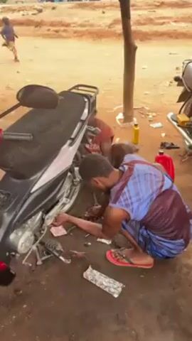 The video shows two men repairing a motorcycle in an outdoor setting. One man, wearing a blue and maroon patterned shirt and red sandals, is kneeling next to the motorcycle and using tools to work on the engine. Another man, wearing a red shirt, is partially visible, assisting with the repair. The motorcycle is black and silver, and there are tools and parts scattered on the ground around it. A bottle of oil and a red container are also visible nearby. In the background, a child is running across a dirt area. A tree trunk stands near the motorcycle. Another motorcycle is parked nearby. The ground is dirt.
