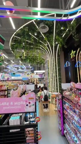 The video shows the interior of a brightly lit store. The ceiling features a decorative canopy made of artificial greenery and white LED strip lights, shaped like a tree. Colorful light tubes curve across the ceiling, and several whimsical, character-themed decorations are suspended from above, including a green cube with a red face and a red character with a white hat. In the foreground, aisles of merchandise are visible, with shelves stocked with colorful products, including what appear to be snacks and personal care items. Several shoppers are present in the store, browsing the aisles and walking through the space. One person is seated at a small table in the background, near the decorated canopy. The overall atmosphere is vibrant and playful, suggesting a retail environment geared towards a younger demographic or those seeking a fun shopping experience.