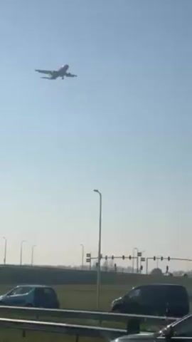 The video shows a DHL airplane approaching for landing under a clear blue sky. The camera appears to be positioned next to a road with guardrails, with traffic moving in both directions. Streetlights are visible along the side of the road.