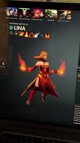 The video captures a computer screen displaying the character selection and strategy phase of the video game Dota 2. The main subject is the game interface, centered on the hero Lina, a female character with orange hair and fiery hands, wearing a red and gold outfit. She is shown floating and performing idle animations.

The activity taking place is a player preparing for a match, specifically during the "STRATEGY TIME," which counts down from 0:13 to 0:10 in the video. The player is interacting with the game's user interface, selecting starting items and potentially a lane. The "Strategy Map" shows "BOTTOM LANE (Dire Offlane)" highlighted. Various inventory items like Magic Wand, Boots of Speed, Null Talisman, Faerie Fire, and Observer Ward are visible, with some marked as purchased and others as recommended.

Notable details include the game being Dota 2, with several hero portraits visible at the top, some with humorous or offensive Russian usernames (e.g., "ЖОПА ЕБАНУТАЯ", "АБАССАБЛЯДЬ", "DRODIK"). A text box at the bottom left lists heroes that have been banned, such as Axe, Omniknight, Techies, Ogre Magi, Lycan, and Huskar. It also states that Pudge was chosen by both teams. The video is filmed from a slightly elevated angle, showing a portion of a laptop keyboard in the foreground, suggesting the game is being played on a portable computer. Reflections are visible on the screen, indicating an indoor setting. This video was filmed in Graz, Austria.