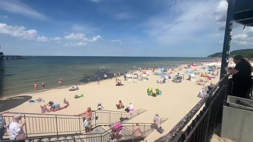 The video shows a wide view of a crowded beach on a sunny day with a blue sky and scattered clouds. The camera is positioned on a set of stairs looking down at the beach and out towards the sea. A pier extends into the water on the left side of the frame. The sandy beach is filled with people sunbathing, sitting under umbrellas, and walking along the shore. Some people are in the shallow water. On the stairs in the foreground, several people are descending or standing, including children. A person in a black shirt is visible on the right side of the frame, looking at their phone. There is a railing in the foreground and a structure on the right that partially obstructs the view.