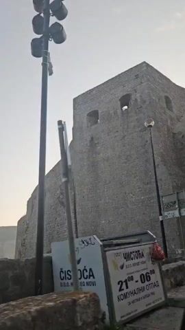 The video pans across a stone fortification with narrow openings. A tall lamppost with multiple lights is visible in the foreground, along with another lamppost with a single light further back. Trees and foliage are present on the left side of the screen. A sign with the word "ČISTOĆA" and a logo is also visible. The sky is overcast. The camera appears to be moving, possibly from inside a vehicle.