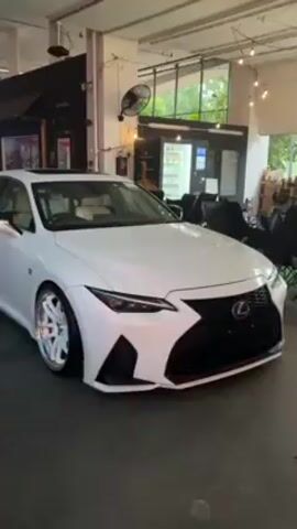 A white Lexus IS sedan with a custom body kit and wheels is shown in a parking garage. The car has a sunroof and beige interior. The wheels are white with red brake calipers, and appear to have a chrome lip. The car is modified with a low ride height. The environment is a brightly lit indoor garage setting with visible vending machines, seating, and other businesses in the background. No individuals are clearly visible in the video. The camera pans slowly around the vehicle to show the modifications.