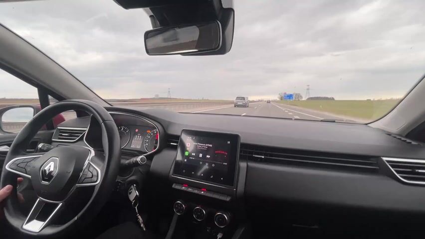 The video shows the interior of a moving Renault car, presumably the driver's perspective. The car is driving on a highway with at least two lanes in each direction, with other cars visible on the road. The sky is overcast and cloudy. On the right side of the highway, there's a green field. Power lines run parallel to the highway. The car's dashboard is visible, showing the steering wheel, instrument panel, and a central touchscreen displaying music from Spotify. The speed of the car fluctuates between 108 and 110 km/h. The driver's hand is visible on the steering wheel. A signpost indicating Minsk is visible in the distance.