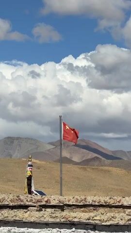 A red Chinese flag with five yellow stars waves gently on a flagpole in a vast, arid, mountainous landscape. To the left of the flagpole stands a colorful chorten-like structure, possibly a stupa, adorned with prayer flags or fabrics. In the immediate foreground, there is a stone wall or barrier. The sky is partly cloudy with large white cumulus clouds set against patches of blue sky, indicating a bright daytime setting. Mountains with varied hues of brown and grey dominate the background, suggesting a high-altitude region. The overall scene depicts a remote, possibly high-altitude, area within China, consistent with Labrang township.