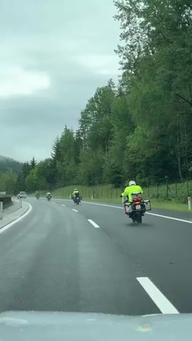 A group of motorcyclists wearing neon yellow jackets ride in single file on a wet, paved highway. The motorcyclists are spaced out with one motorcycle visible in the foreground and several others further down the road. The highway is surrounded by a dense forest with green trees. The sky is overcast and appears to be cloudy. The perspective suggests the video is filmed from another vehicle on the highway.