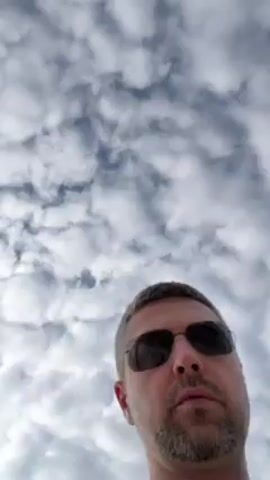 A man wearing sunglasses looks up at the camera against a backdrop of a cloudy sky. The man has a gray beard and graying hair. The camera is positioned low, looking upwards.