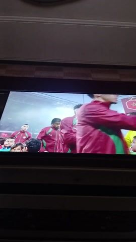 The video shows a television screen displaying footage of a group of men, presumably athletes, wearing matching burgundy tracksuits with green accents. The back of one tracksuit reads "المغرب" in Arabic, which translates to "Morocco." The athletes are greeting each other, possibly before or after a game, with handshakes and smiles. The environment appears to be a stadium or arena with bright lighting. The television is set in a home setting with a decorative ceiling.