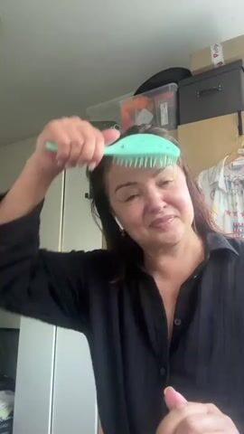The video features a middle-aged woman in Steenwijk, Netherlands, in an indoor setting that appears to be a bedroom or dressing area. She has brown hair and is wearing a black shirt and white earbuds. The woman is holding a light green hairbrush and is in the process of brushing her hair. She is smiling and seems to be in a good mood, possibly recording a video for social media. Behind her, there are shelves with boxes and clothing. The lighting suggests it is daytime.