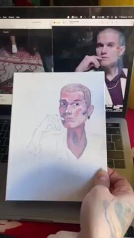 A hand holds a small, rectangular canvas with a partially completed portrait. The portrait depicts a bald or short-haired person with visible facial features rendered in shades of red, purple, and pink. The person is wearing a white shirt, the outline of which is sketched in pencil. The background of the canvas is plain white. Behind the canvas, a laptop is open, displaying two screens. On the left side of the laptop screen, a YouTube video is playing, showing a group of people on a brightly colored rug. On the right side, another image displays the person from the portrait, also a YouTube video. The subject is wearing a purple shirt with a white jacket and a gold chain. The laptop is sitting on a red surface, presumably a table. The hand holding the canvas has a tattoo. The overall lighting is bright and even.