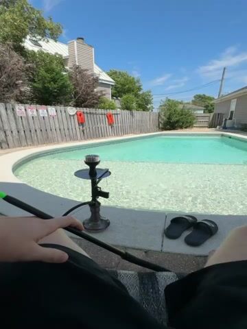 A person is relaxing by a swimming pool on a sunny day. The pool has clear, turquoise water and a shallow lounging area. A hookah is set up on the edge of the pool, and a person's hand is visible holding a hookah hose. A pair of black flip-flops rests on the pool deck. In the background, there is a wooden fence with several red life preservers attached to it, and a residential building with grey siding and white trim. The sky is blue with a few scattered clouds. The time of day appears to be midday, and the weather is warm and pleasant. The overall scene evokes a sense of leisure and relaxation.