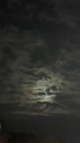 The video shows a cloudy night sky with the moon partially visible through breaks in the clouds. The moon is illuminated and casts a soft glow. In the foreground, the silhouette of a building with a green roof is visible, along with some trees. The overall mood is dark and somewhat eerie.