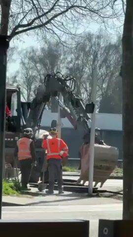 The video shows construction workers digging near a street. An excavator with a large bucket is visible, and two workers in orange vests are standing nearby. One worker is holding a shovel. A traffic light shows a red pedestrian signal. A silver car passes by. The trees in the background are bare. The weather appears sunny.