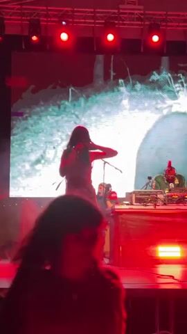A performance is taking place on a stage. A female performer in a dress and boots is singing into a microphone. She has dark hair and is moving her arms. Behind her, a person with a red mohawk and a green jacket is operating DJ equipment. The background features a large screen displaying a grey, abstract image with some red accents. Red stage lights illuminate the scene. In the foreground, the silhouette of a person's head and shoulders is visible, obscuring part of the stage.