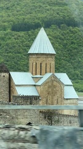 The video shows a stone church with a silver roof, surrounded by a stone wall. The church is set against a backdrop of lush green hills. The camera pans slightly to the left, revealing more of the building and its surroundings. The video appears to have been taken from a moving vehicle.