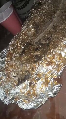 A video shows a close-up of leftover fish bones on aluminum foil. Some small pink cups are also visible in the background. A person says, "As you can see, the fish, the fish don't finish."
