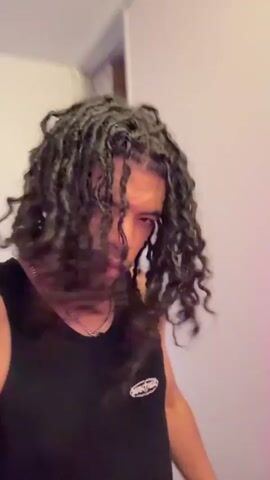 The video shows a medium shot of a man with long, dark hair styled in dreadlocks. He is wearing a black tank top and a silver chain necklace. He has a tattoo on his neck that reads "1974". The background is a white wall with a built-in white shelf unit. The lighting in the room has a pink hue. The man initially looks down, then looks up at the camera and throws his head back, tossing his hair up and back.
