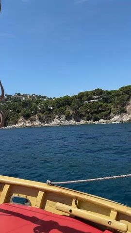 This is a first-person perspective video taken from a wooden sailboat navigating along a coastline. The water is a deep blue and shows gentle ripples, indicating a calm sea. The sky is clear and a bright blue, suggesting a sunny day.

In the foreground, the wooden deck of the sailboat is visible, with a red cushioned seat. Ropes are coiled and attached to the mast, which is partially visible and made of a light-colored material.

The background features a verdant, hilly coastline dotted with houses and trees. The terrain is rugged with rocky outcrops leading down to the sea. In the distance, several sailboats and yachts are anchored or sailing, adding to the nautical scene.

The overall impression is one of a leisurely sailing trip on a beautiful, sunny day, likely in a coastal region known for its scenic beauty and sailing opportunities. The location is identified as Palafrugell, Spain, which aligns with the visual cues of the Mediterranean coastline. The time of day appears to be midday, given the bright sunlight. There is no visible text in the video.