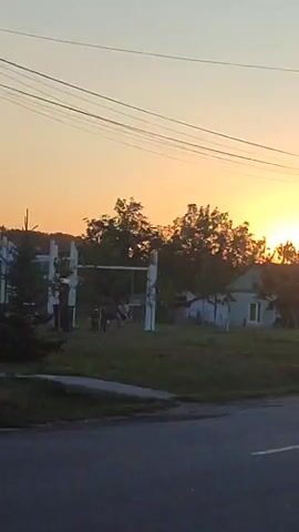 The video shows a sunset in a rural setting. A swing set is visible in the foreground, with a few people standing around it. A house is situated behind the swing set, and trees surround the area. Power lines are strung across the top of the frame. The sun is low on the horizon, casting a warm glow over the scene.