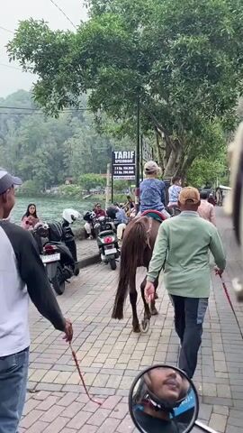 The video depicts a scene on a cloudy day near a lake. In the foreground, a person is riding a horse on a brick-paved walkway. Several people are sitting or walking along the walkway, some by the water's edge. Several scooters are parked nearby. A sign displays the "TARIF SPEEDBOAT" prices. The background shows the lake with mountains covered in trees and some mist. The video appears to be taken from the perspective of someone on a motorcycle, judging from the mirror view and motion.