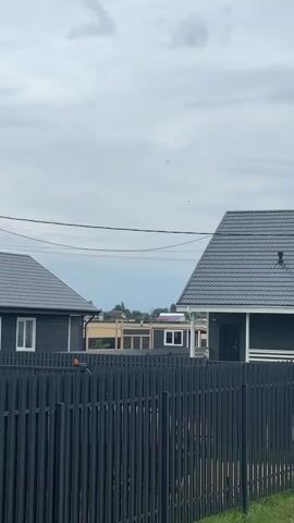 The video shows two paragliders in the sky above a suburban area with houses and a fence. The sky is cloudy and the overall lighting is overcast. The paragliders are visible as small figures against the sky, with one paraglider appearing to fly behind a house. The video is shot from a low angle looking up at the sky.