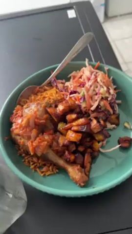 The video shows a plate of food. The plate is green and contains jollof rice, chicken, plantains, and coleslaw. A spoon rests on top of the jollof rice and chicken. The plate is sitting on a black surface.