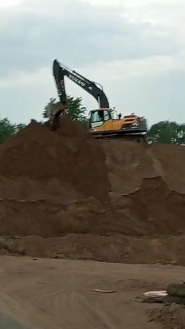 The video shows a yellow and black excavator working on top of a large pile of dirt. The excavator, identified as a Volvo by the lettering on its arm, is positioned on the peak of the dirt pile with its boom and bucket extended. The bucket is filled with dirt. The background includes trees and an overcast sky. The ground in the foreground is also dirt.