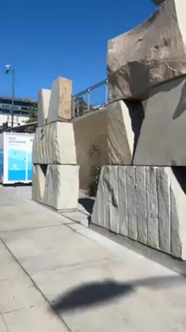 The video shows an exterior shot of a concrete sidewalk and adjacent street. On the left, a large, rough stone structure stands adjacent to the sidewalk. It appears to be constructed of large, irregularly shaped stone blocks stacked on top of one another. Behind the structure, at a slightly higher level, is a metal railing, suggesting a raised platform or walkway.  Behind the stones are growing shrubs with white flowers. The sidewalk runs parallel to the street. The street is divided with a double white line in the center. On the opposite side of the street are several yellow barriers and a bike lane symbol painted on the road. Across the street are buildings and palm trees. The sky is clear and blue. Street lights are visible along the sidewalk.  The video appears to be taken while moving or panning slightly to the right, providing a wider view of the scene.