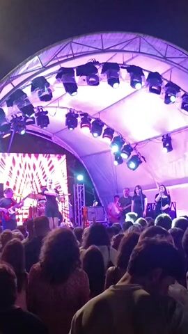 The video shows a band performing on a stage under a large tent structure. The stage is lit with purple and white lights, and a large screen displays a geometric pattern. There are several band members on stage, including a singer and a guitarist. A crowd of people is gathered in front of the stage, watching the performance. The video is taken from behind the crowd, looking towards the stage. The environment appears to be outdoors, with trees visible in the background. The time of day is likely evening or night, given the lighting.