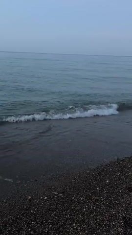 The video shows a peaceful beach scene with gentle waves lapping the shore. The water is a muted blue-green, and the sky is a soft, pale blue with a few wispy clouds. The beach is made of dark pebbles and sand, with some footprints visible. In the distance, a wooden pier extends into the water. The overall atmosphere is tranquil and serene, with the sounds of the waves and the sea birds creating a natural soundtrack.